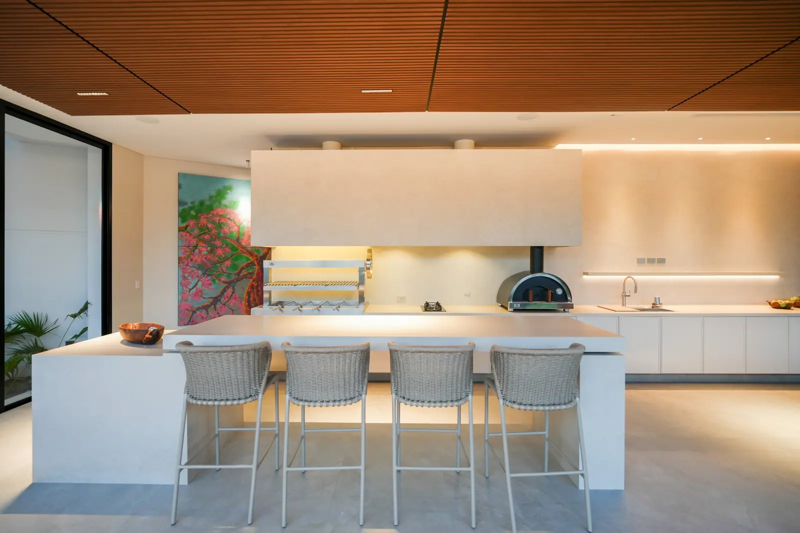 Área social de la Casa AW con la cocina integrada: churrasquera y horno Averno sobre una mesada clara, isla con cuatro banquetas y un mural, bajo un cielorraso de listones de madera