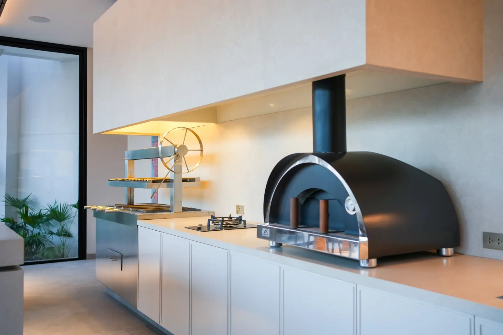 Horno Averno negro con chimenea junto a una churrasquera con volante y un anafe, sobre la mesada de la Casa AW