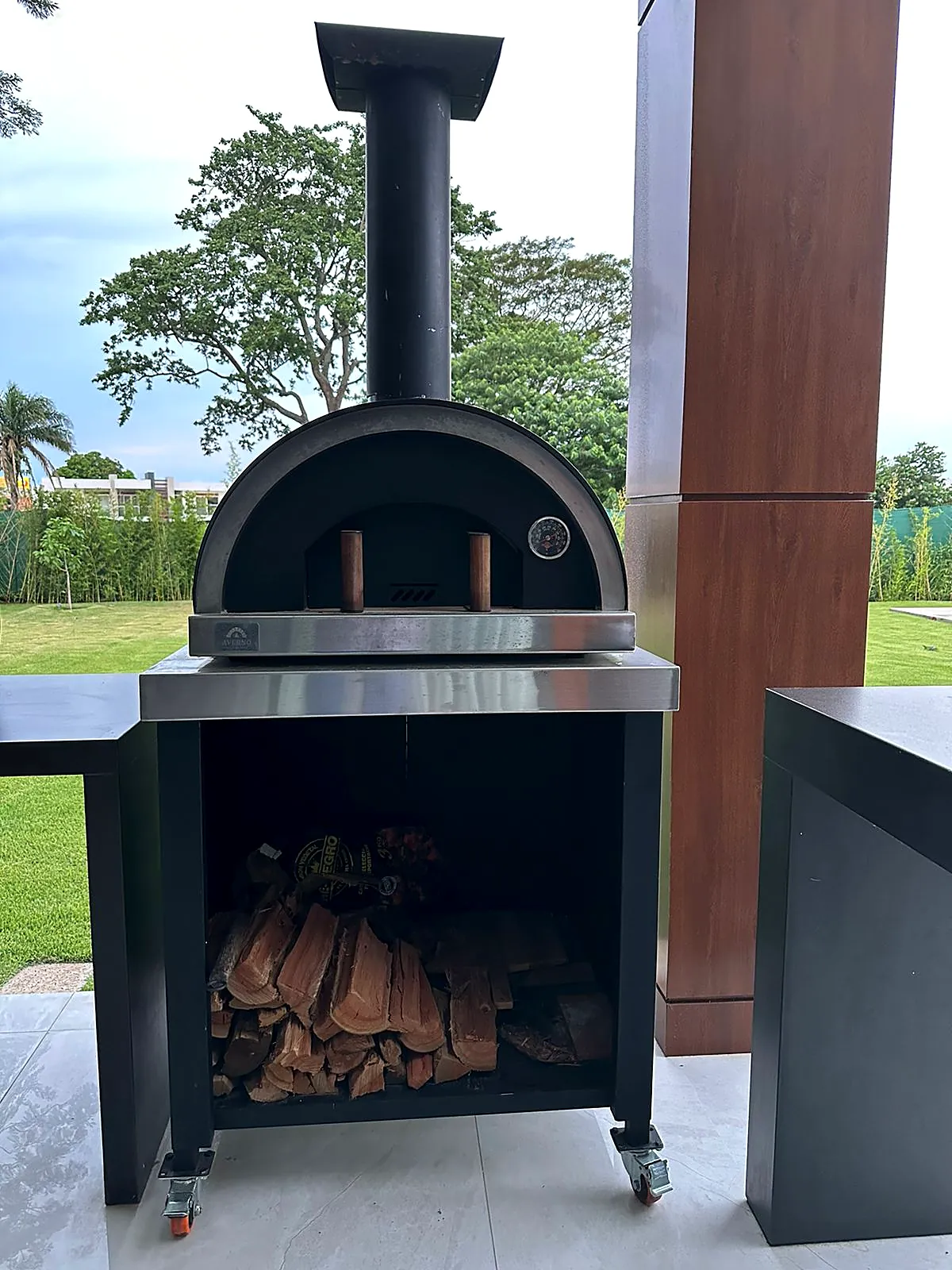 Horno Averno negro con chimenea sobre una base de acero con ruedas y espacio para guardar leña