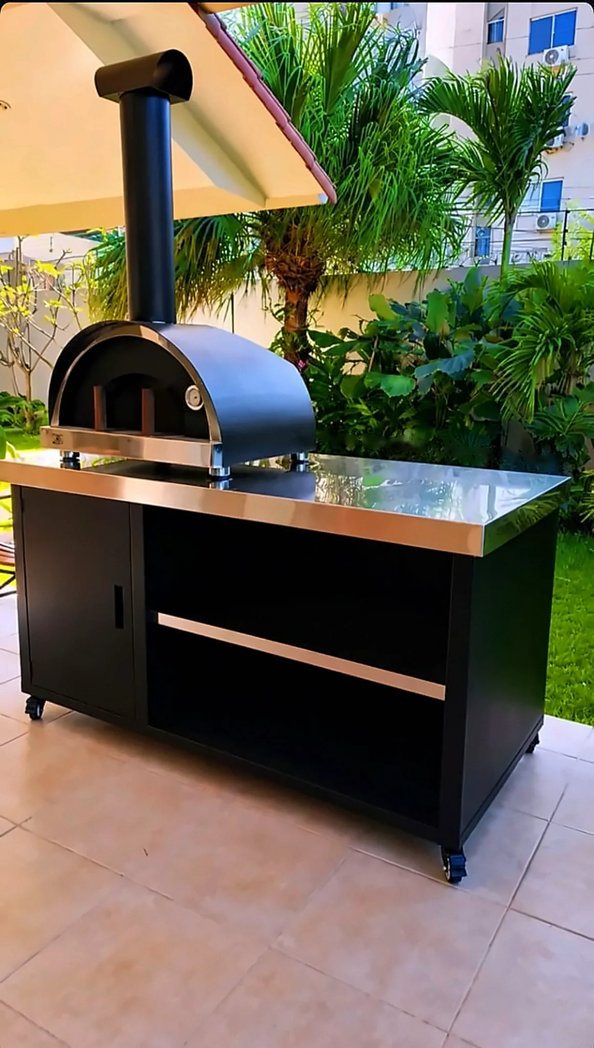 Base movible negra con tapa de acero, puerta y estante, con un horno Firenze encima, en una terraza con plantas