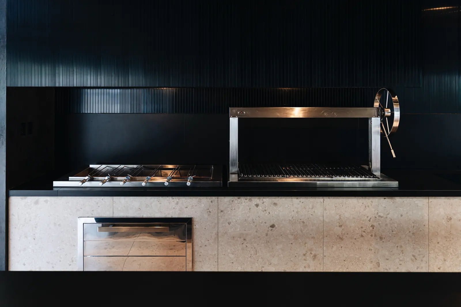 Cocina del club house con brochetero y churrasquera Averno sobre una mesada negra, frente a un muro oscuro