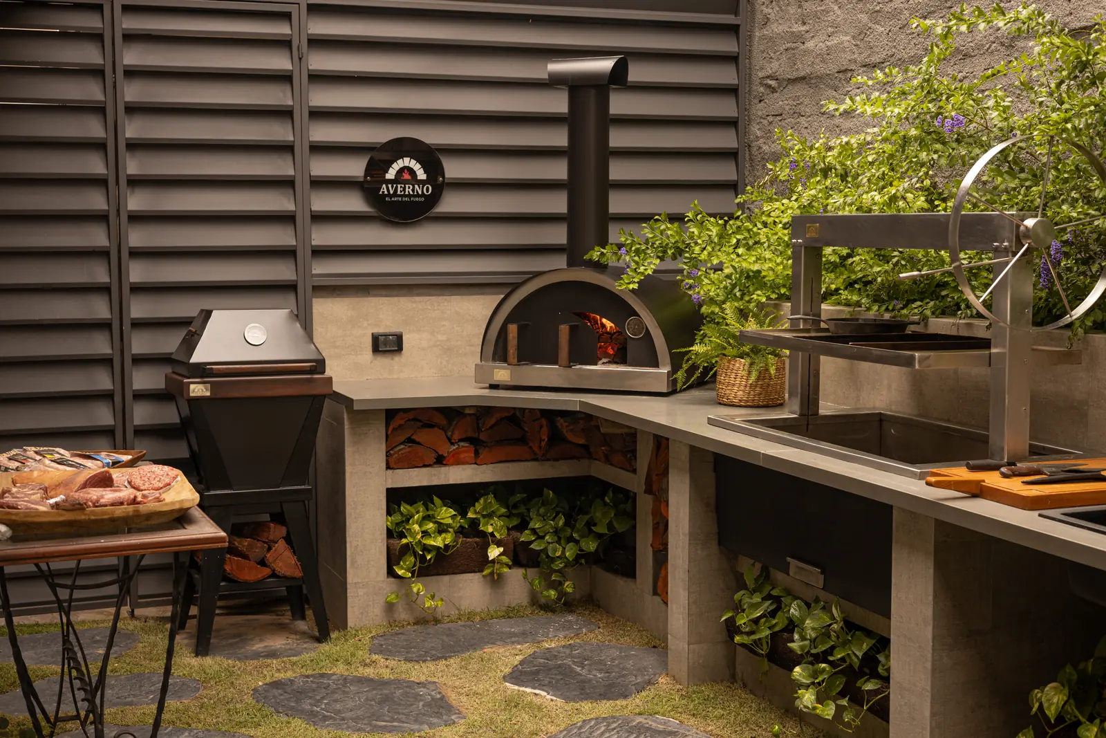 Horno Averno encendido junto a una churrasquera con volante, entre plantas, en el showroom de Cronembold