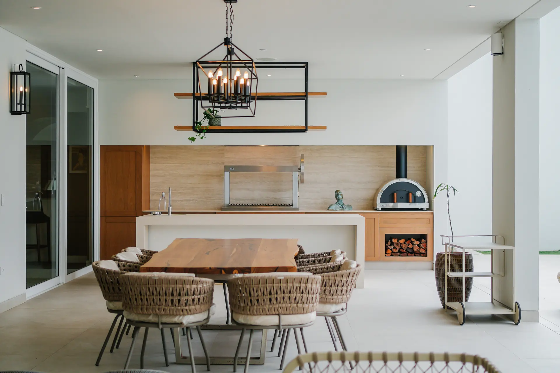 Cocina exterior con churrasquera y horno Averno integrados a un mueble de travertino y madera, junto a un comedor de mesa maciza