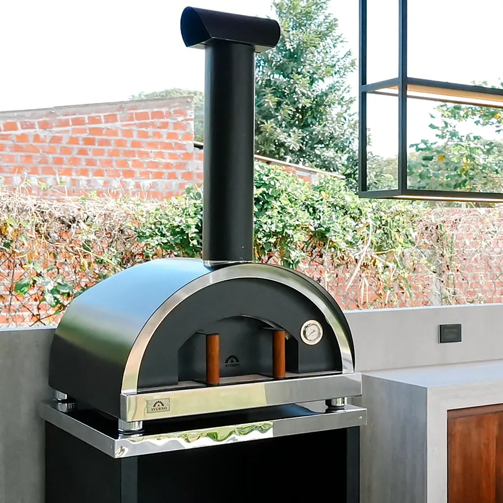 Horno Averno Firenze XL negro con chimenea y cinta de acero, sobre una base negra, junto a una isla de hormigón y madera
