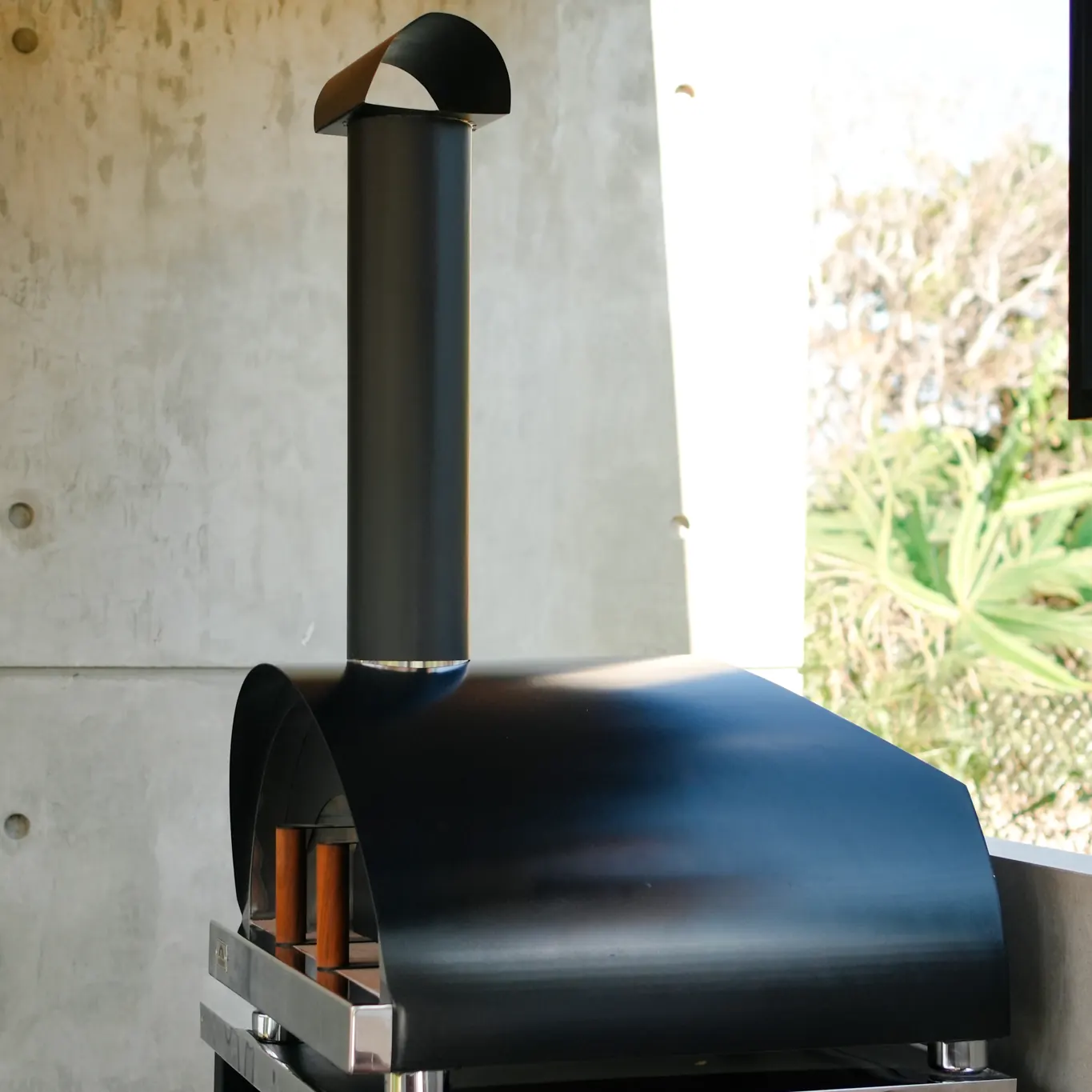 Horno Averno Firenze XL negro de perfil, con su chimenea y sombrerete, frente a un muro de hormigón
