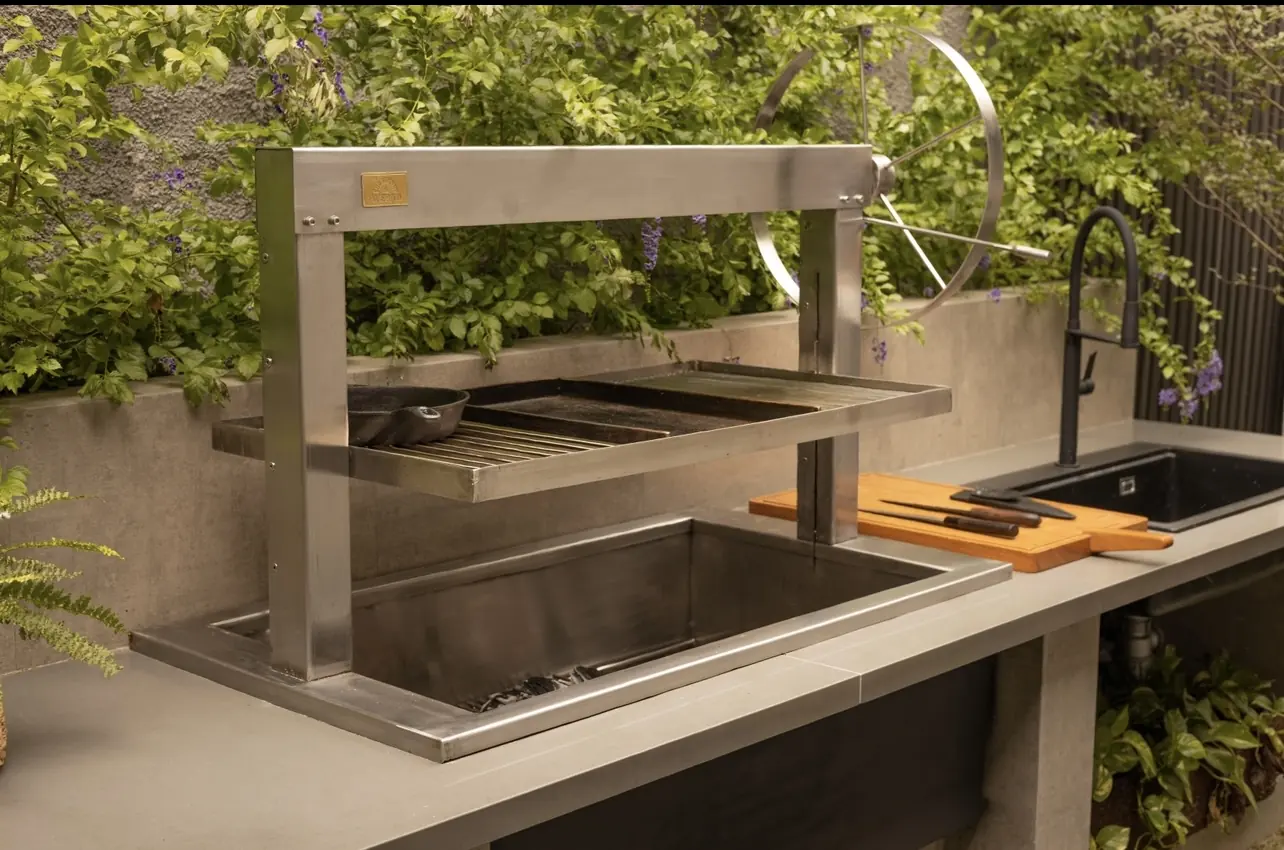 Churrasquera Averno con volante y parrilla elevada sobre una mesada rodeada de plantas