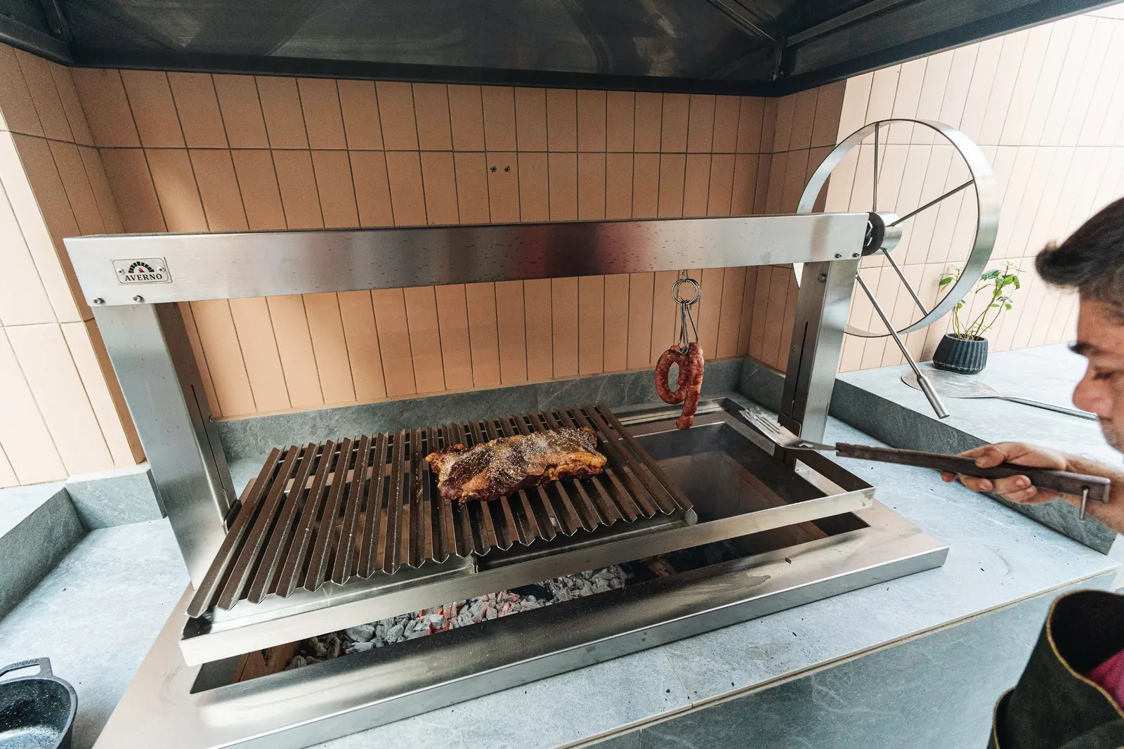 Churrasquera Averno con carne sobre la parrilla y brasas, con volante de acero, en el Showroom Tumpar